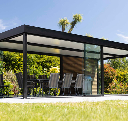 Strakke aluminium tuinkamer met glazen schuifwanden, ingericht met eettafel en barbecue in een zonnige tuin